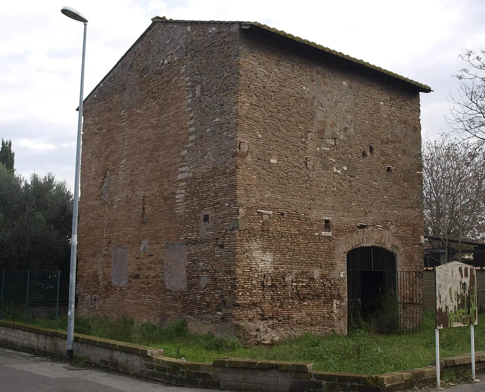 A plain, box-like brick building with a pitched roof and a barred arched doorway, standing behind a low wall beside a street lamp.