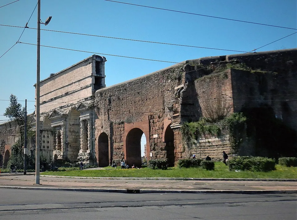 A massive gate of pale stone with a tall plain attic above its arches, joined on the right to a long stretch of brick wall with an arched opening, beside a road with overhead wires.