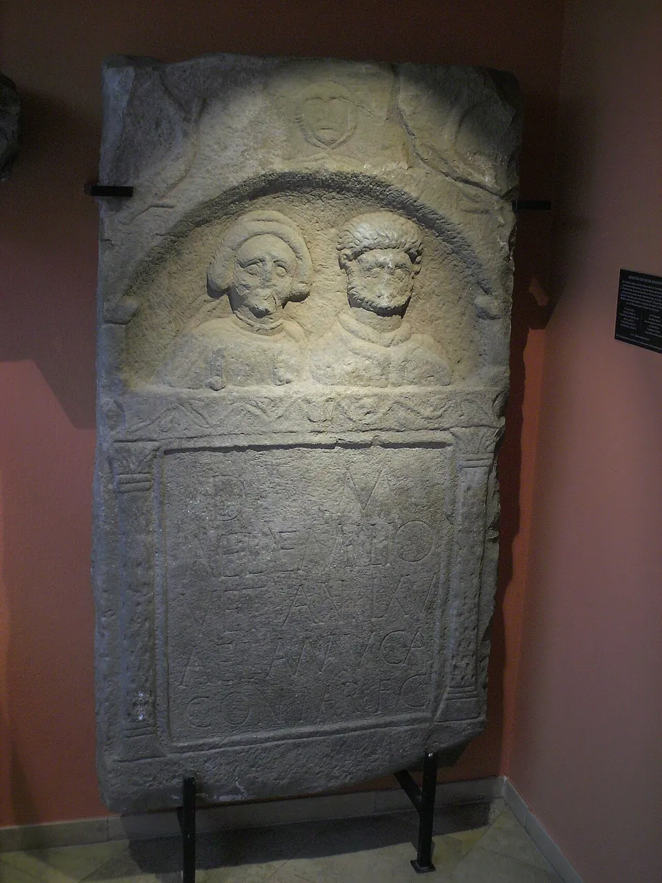 A tall carved gravestone with two portrait heads in a round-arched niche above a panel of inscription, standing against a red wall.