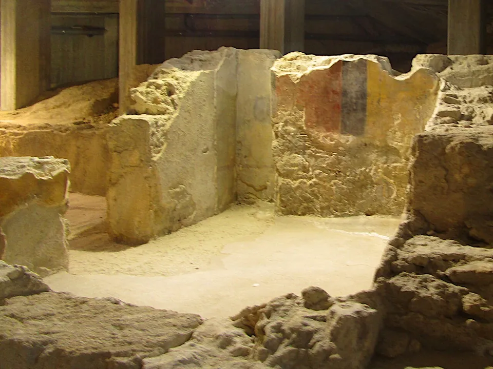 Inside an excavated house, low walls of pale stone, one carrying a patch of painted plaster in vertical bands of red and dark blue, under timber roof beams.