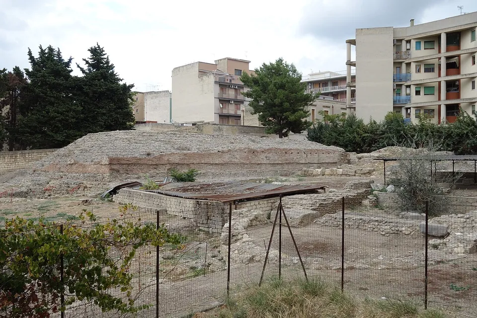 A long, low podium of rubble and brick inside a fenced excavation, a sheet-metal shelter over one part, with pine trees and modern apartment blocks close behind.