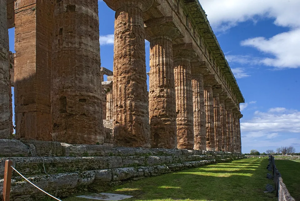 Looking along the side of a Greek Doric temple, a row of massive fluted columns of weathered honey-coloured stone carrying stone beams, standing on a stepped base beside a mown lawn under a blue sky.