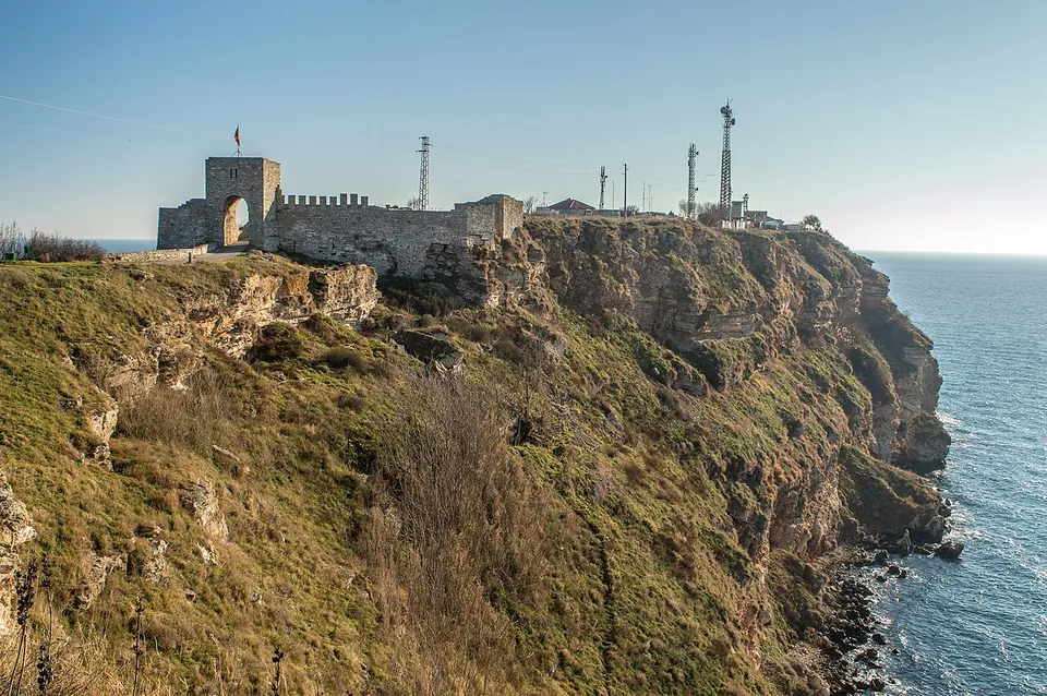 A fortress gate tower with an arched opening and a stretch of crenellated wall stand on the edge of a grassy headland of reddish cliffs dropping into the sea, with radio masts behind.