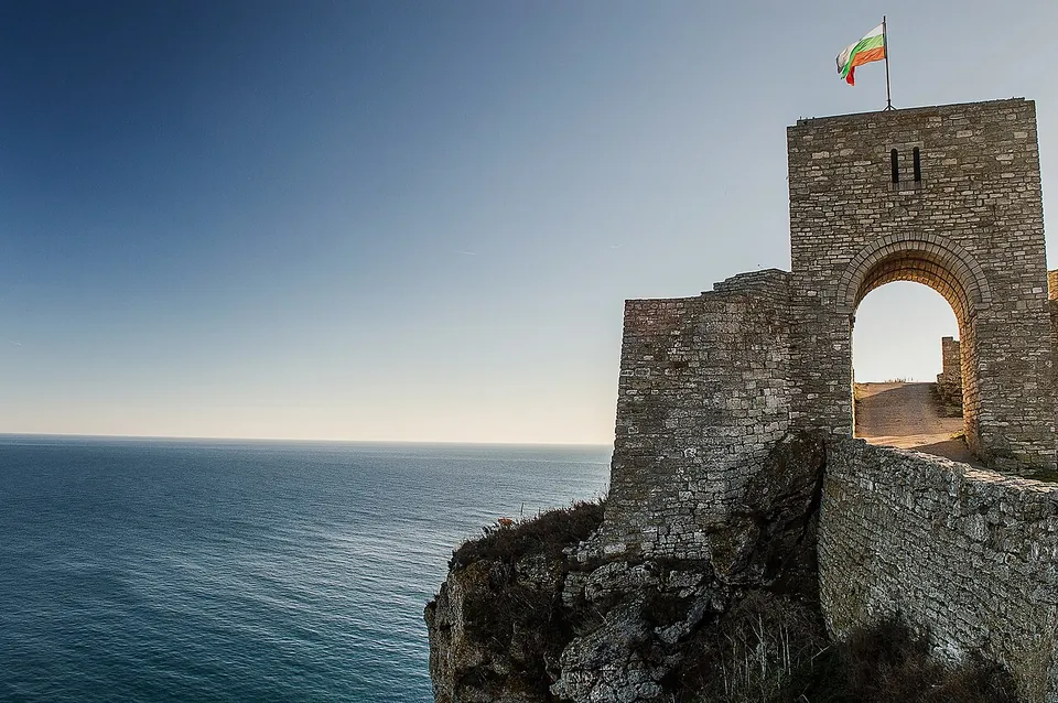 A square stone gate tower with a round-arched passage and a flag on top, joined to a stone wall running along the cliff edge, with the open sea on the left.