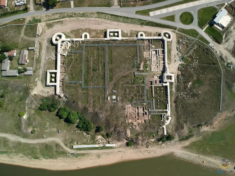 An aerial view of a rectangular fortress on a bluff above a wide river, its walls and white-topped towers outlining the enclosure, with excavated foundations inside, a road and a few buildings beyond.