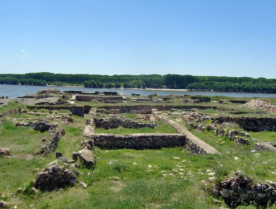 Ruined stone walls and the foundations of rooms in green grass on a terrace, with the broad Danube and its wooded far bank behind under a clear blue sky.