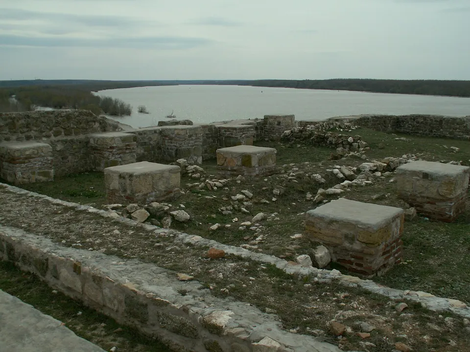 A walkway along the top of a stone wall in the foreground, with square brick pillars standing on the excavated floor below and the wide river under a grey sky behind.