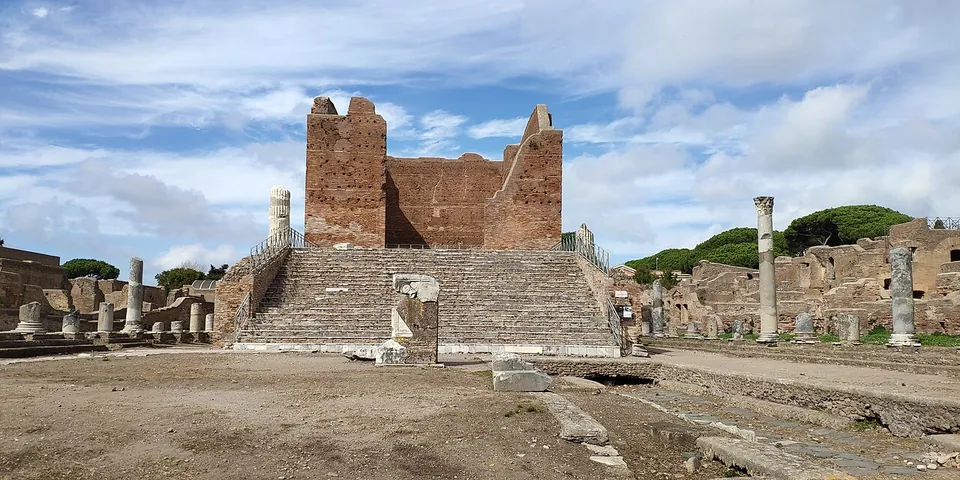 A tall roofless temple of red brick standing on a high podium at the top of a broad flight of steps, at the end of an open square with a few columns and stone fragments.