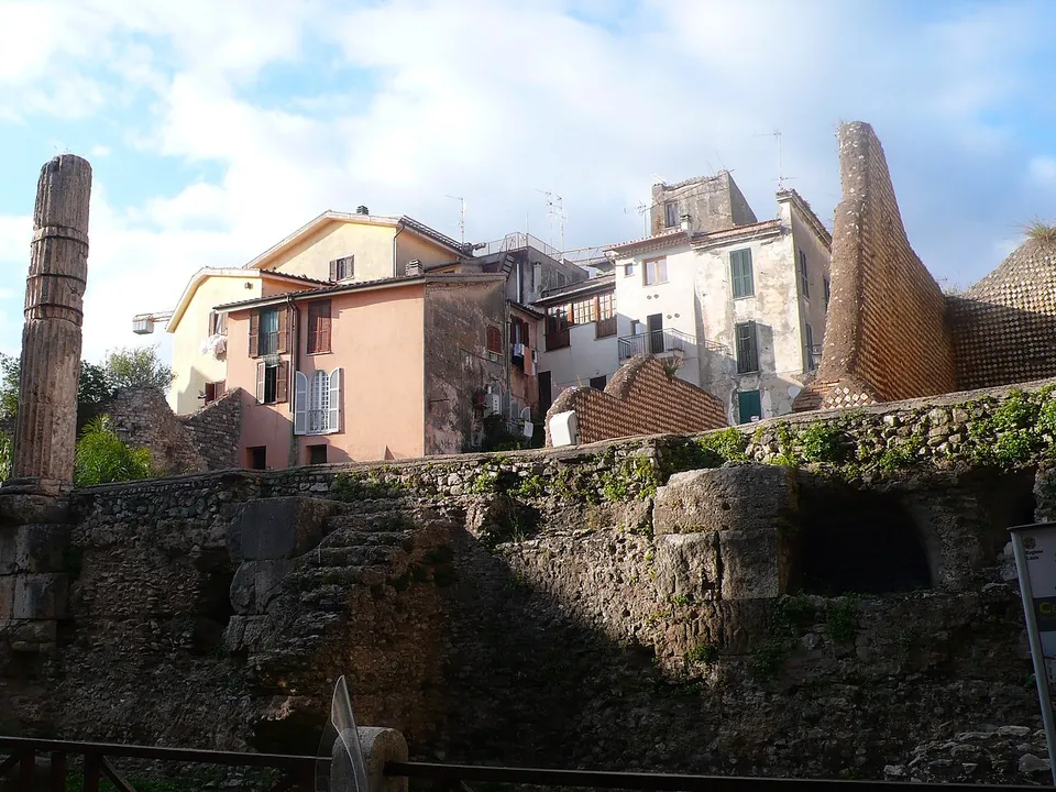 A single tall fluted stone column standing at the left end of a weathered stone podium wall, with a steep triangular fragment of netted-pattern masonry rising behind it among apartment houses.