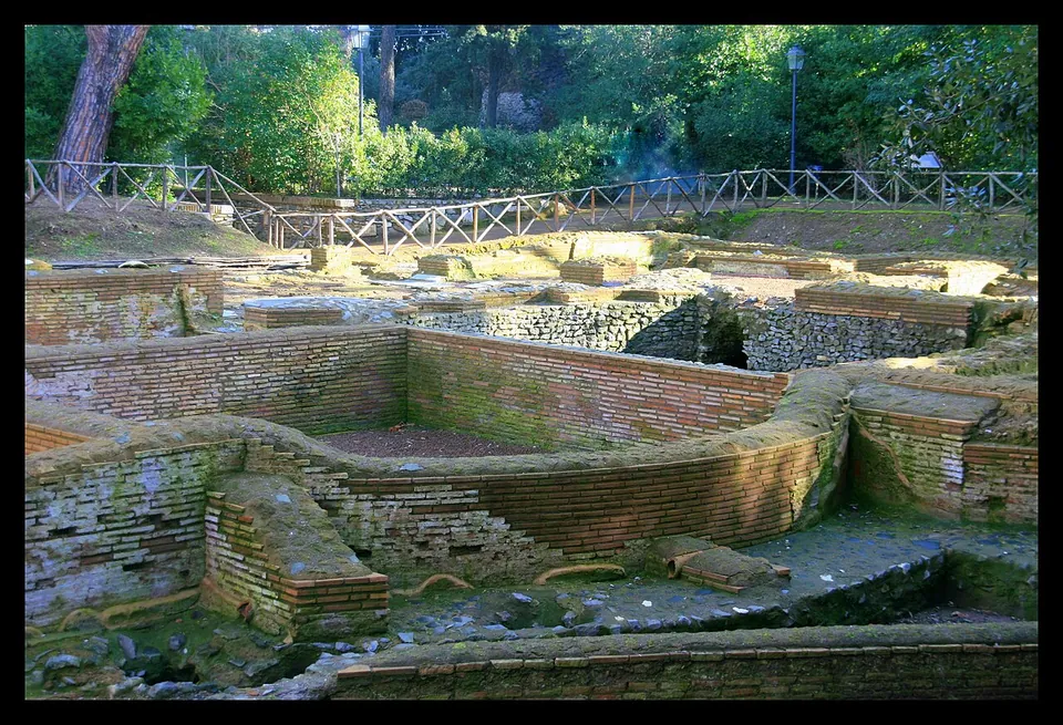 Excavated low brick walls of several rooms, some with curved ends, in a grassy hollow fenced with a wooden rail and backed by trees.