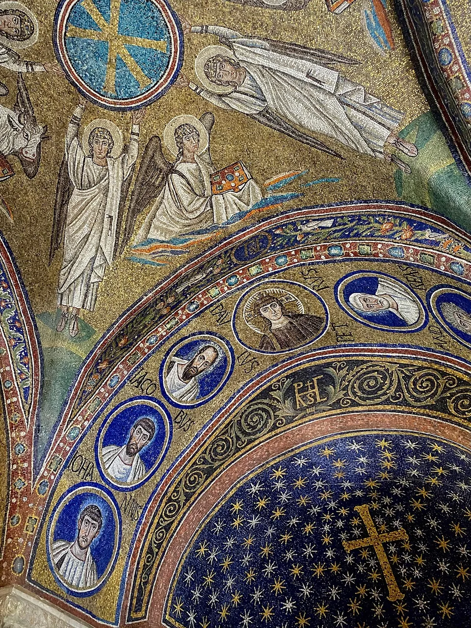 Looking up into a corner of the chapel's vault, covered in gold mosaic with white-robed winged angels; below them an arch lined with portrait medallions on a blue ground frames a small dome of gold stars on deep blue with a gold cross at its centre.