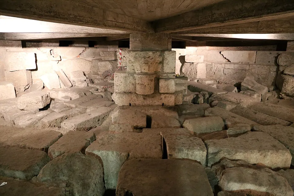 A low underground space lit by lamps, its floor a jumble of large squared stone blocks, with a pier of stacked blocks and a column drum rising to a modern ceiling.