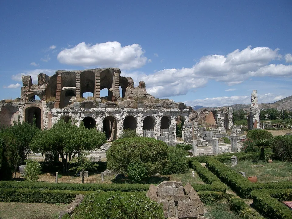 The surviving section of the amphitheatre's outer wall, a row of arches in pale stone with a broken upper storey of brick piers above, behind clipped hedges, trees and scattered stone blocks.