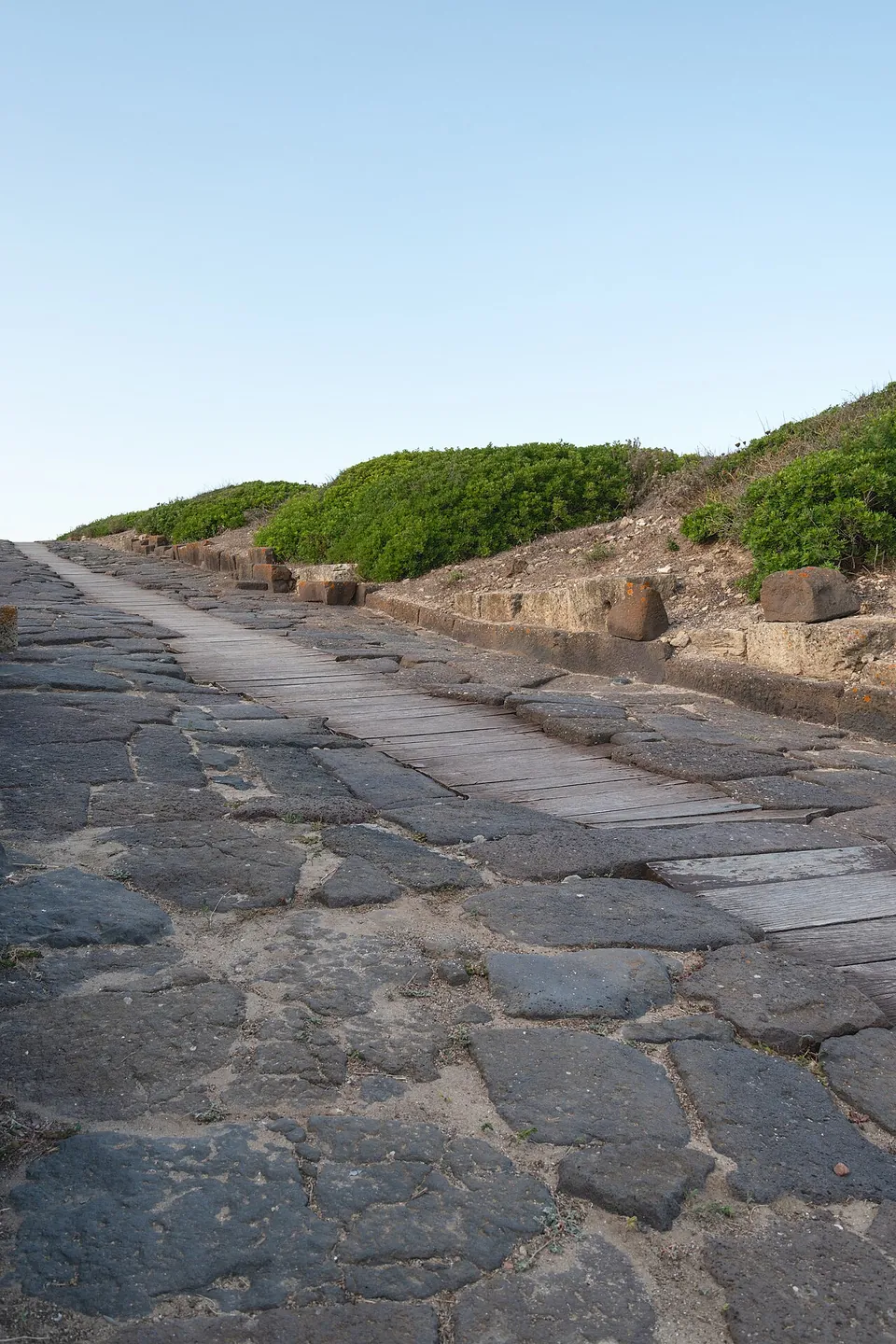 A street paved with large, irregular dark stone slabs rising gently between low stone kerbs and scrub, with a strip of wooden planks laid along part of it.