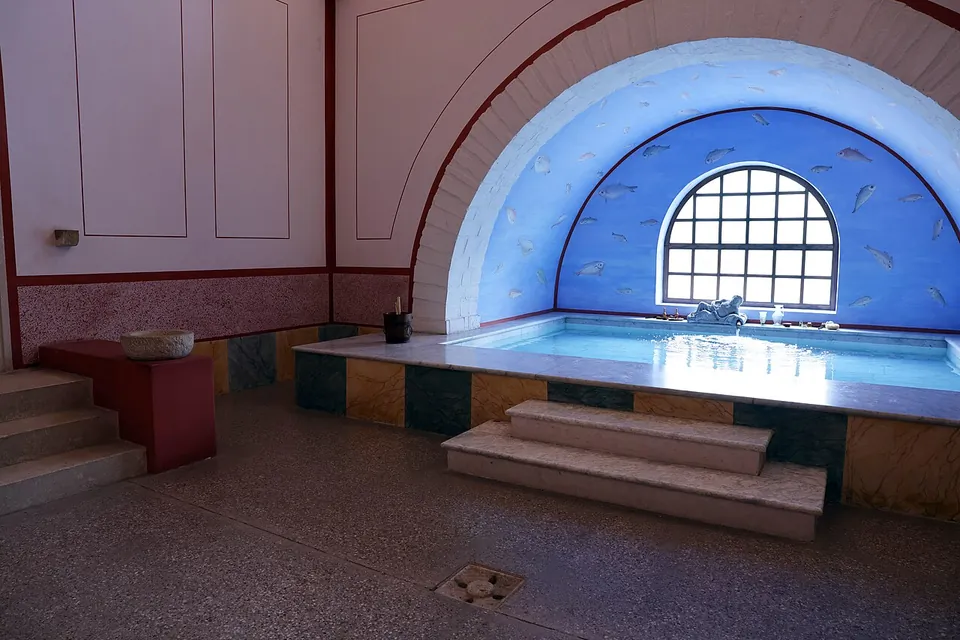 A small bathing pool lined with coloured stone under a blue-painted apse with an arched, gridded window, in a room with red-bordered white walls.