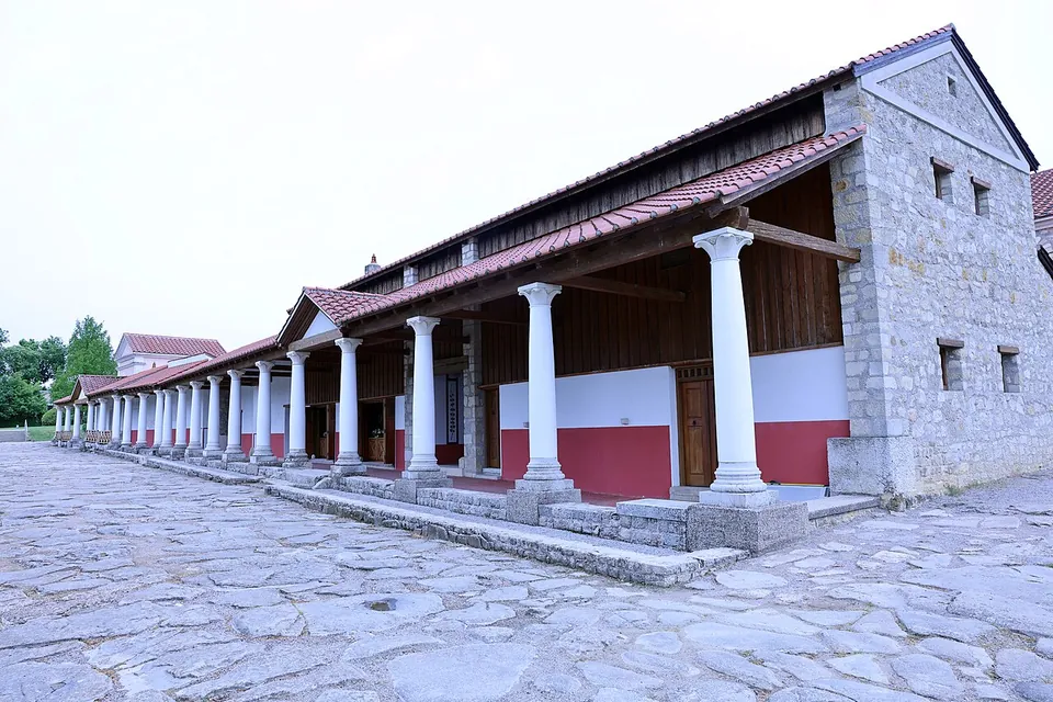 A rebuilt Roman building of grey stone with a long colonnade of white columns under a tiled roof, along a street paved with large irregular stone slabs.