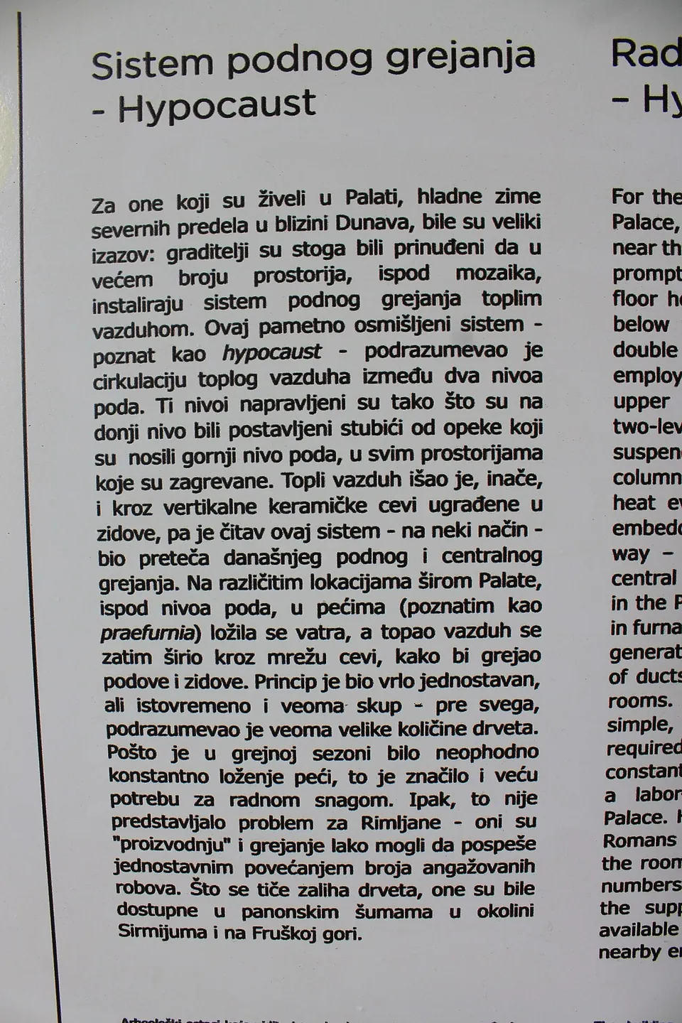 A white interpretation panel headed "Sistem podnog grejanja – Hypocaust", with a full column of Serbian text and, cut off at the right edge, the start of the same text in English.