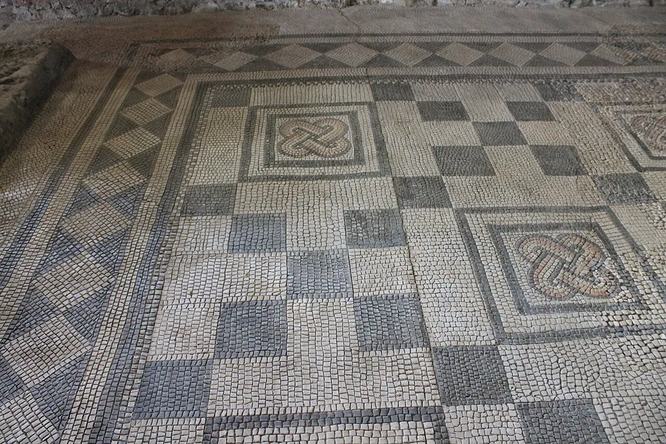 A floor mosaic of small dark and pale stone cubes laid as a chequerboard, with two framed square panels of interlaced knots, one worked in pink and grey, and a band of lozenges along the edge.