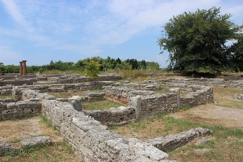 Low walls of fitted grey stone blocks marking out the rooms of an excavated house in rough grass, with two brick column stumps at the left and trees beyond.