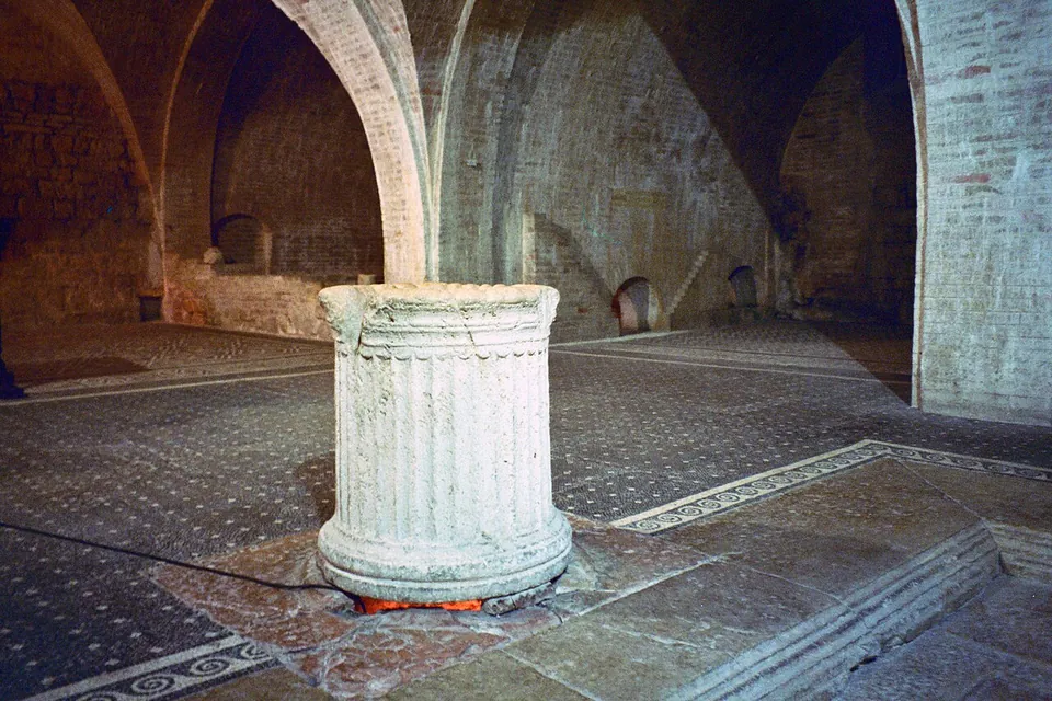 A fluted stone well-head standing on a floor of small dark mosaic tesserae with a patterned border, under low brick vaults lit by spotlights.