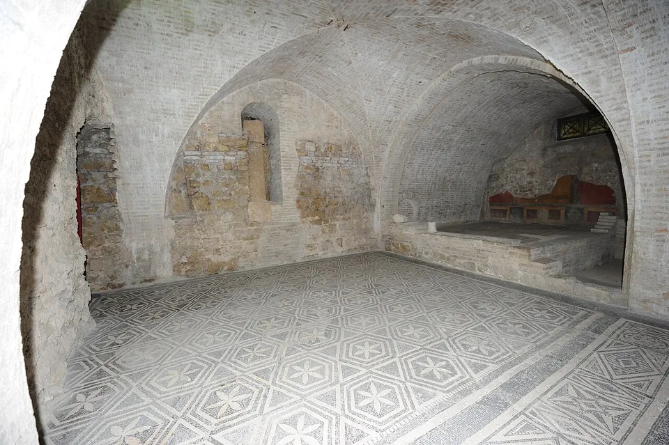A room under a modern brick vault with a white mosaic floor patterned in hexagons and six-petalled flowers, rough stone walls at the back, and a raised floor beyond on the right with traces of red wall painting.