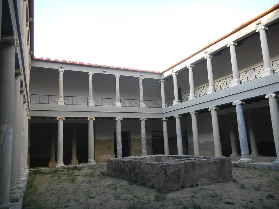 A two-storey courtyard ringed by slender marble columns on both floors, with a railed gallery above and a stone basin in the middle of the earth floor.