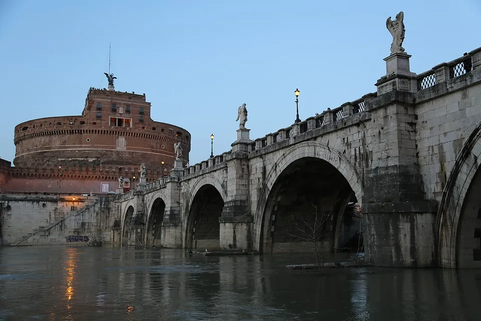 The round castle rises at the left above the river at dusk; a stone bridge of round arches, lined with statues and lamps, crosses the high, still water towards it.