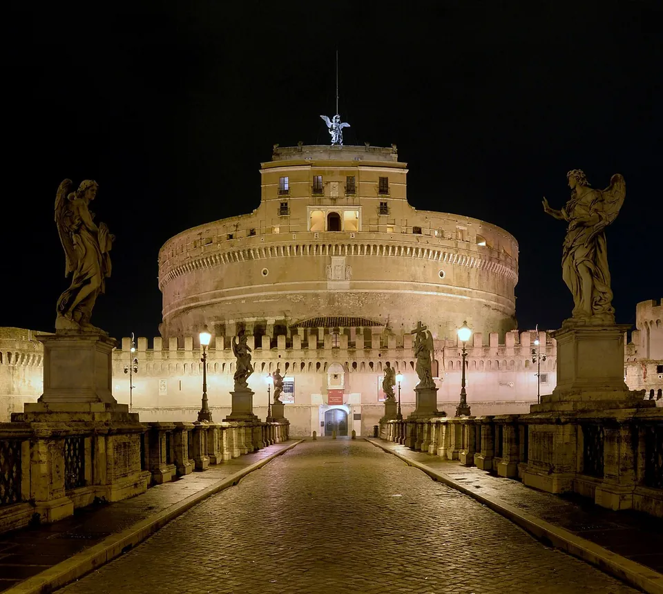 The castle at night, seen straight down the paved bridge, a broad floodlit cylinder with a squarer block and a winged statue on top and crenellated walls at its foot, with statues of angels on tall pedestals along both parapets of the bridge.