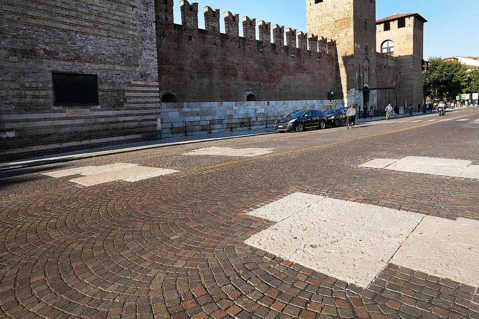 A cobbled street curving past the crenellated brick walls of a castle, with large rectangles of pale stone slabs set into the cobbles in the foreground.