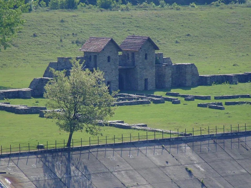 The reconstructed gate and walls of the small fort seen from above, with foundations in the grass and, in the foreground, the concrete slope of a river embankment behind a fence.