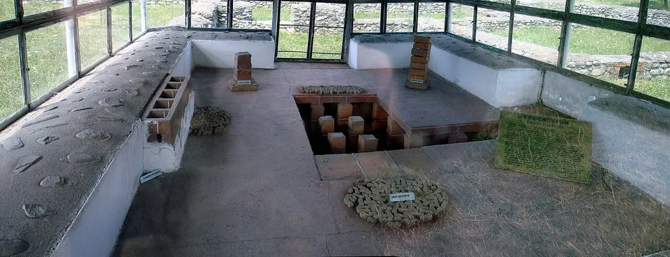 Inside a glazed shelter, rows of small brick stacks stand in a sunken pit below floor level, with low white-rendered walls around it and pieces of stone and brick laid out for display.