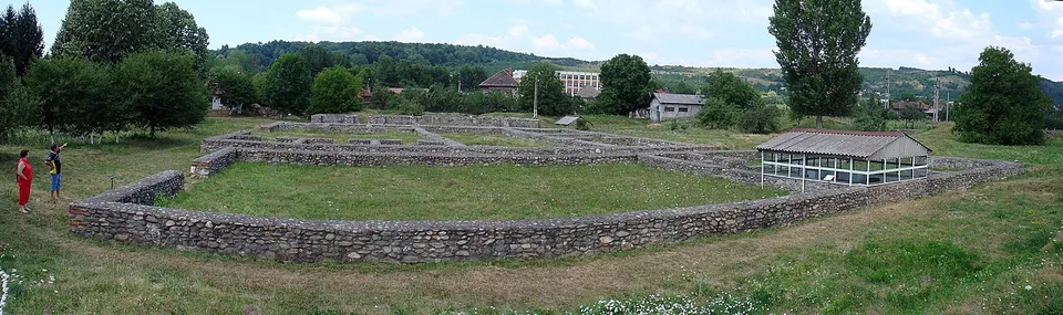 A panorama across a large mown enclosure with the low stone walls of buildings, a long low modern shelter on the right, and houses and wooded hills beyond.