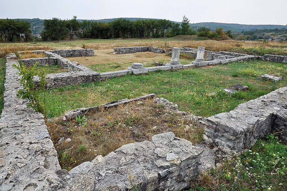 Consolidated stone walls, knee to waist high, outlining rooms in the grass, with short white columns standing in a row behind and hills in the distance.