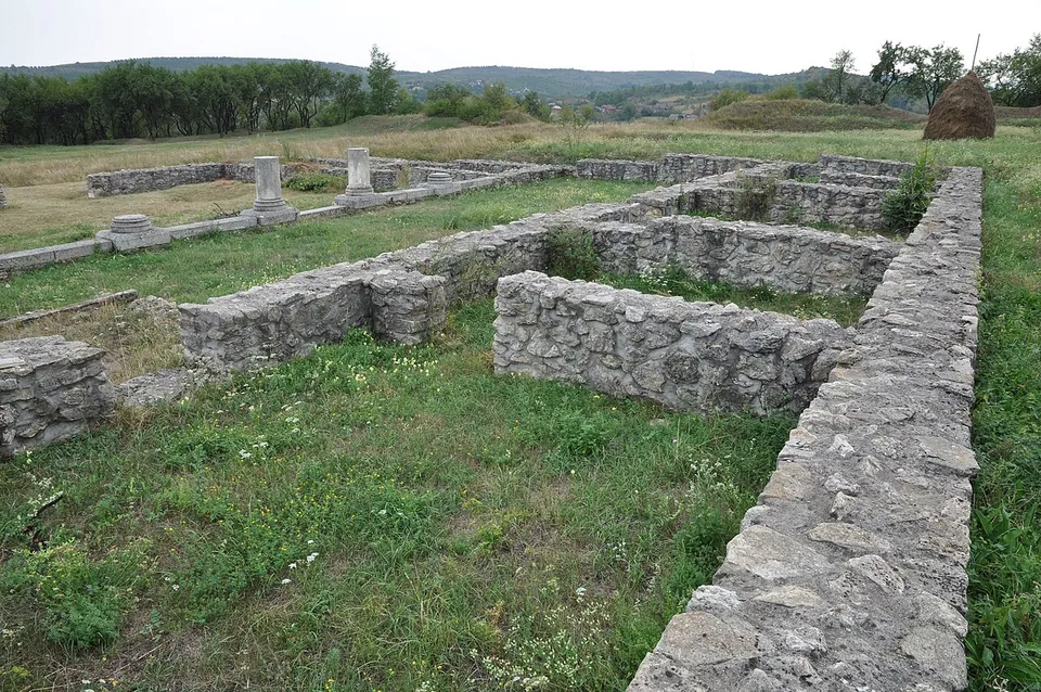Low stone walls of adjoining rooms in green grass, with a haystack and the white column bases of a courtyard in the background.