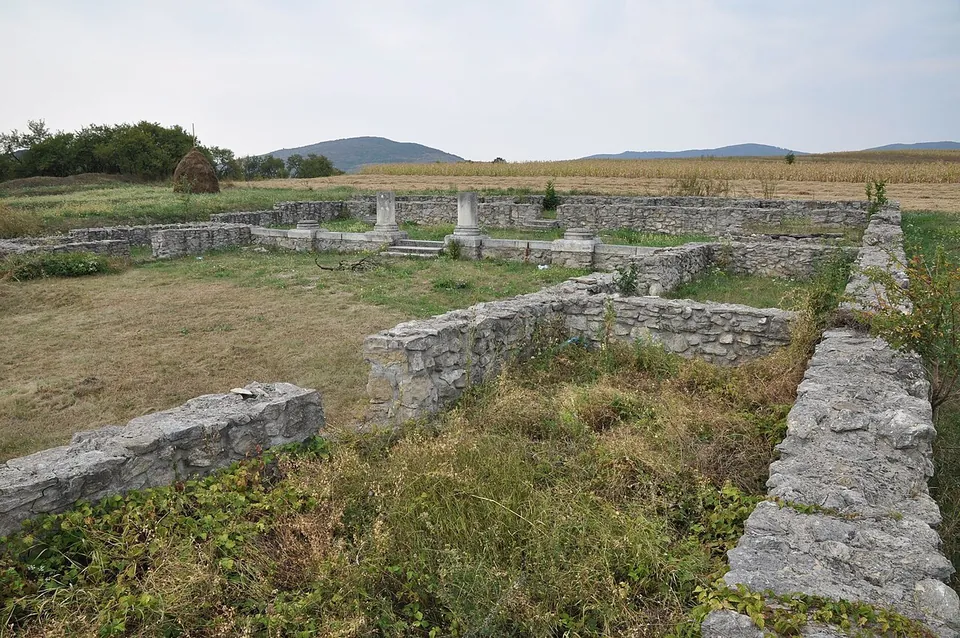 Consolidated low stone walls of an excavated building in rough grass on a gentle slope, a row of short white column bases beyond and wooded hills on the horizon.