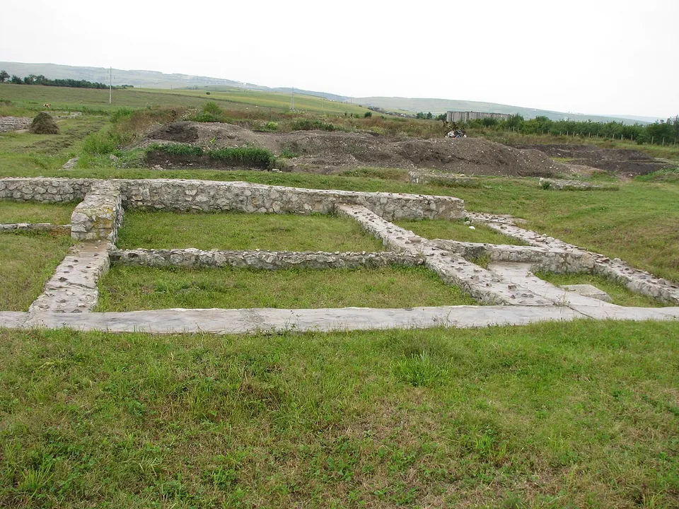 Low consolidated stone walls outlining rooms on an open grassy plateau, with a field and a line of hills on the horizon.