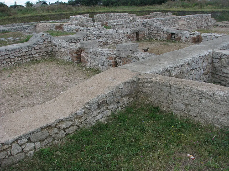 Consolidated stone walls about knee high enclosing rooms with sandy floors, a few short columns of brick and stone standing in them, with grass around.