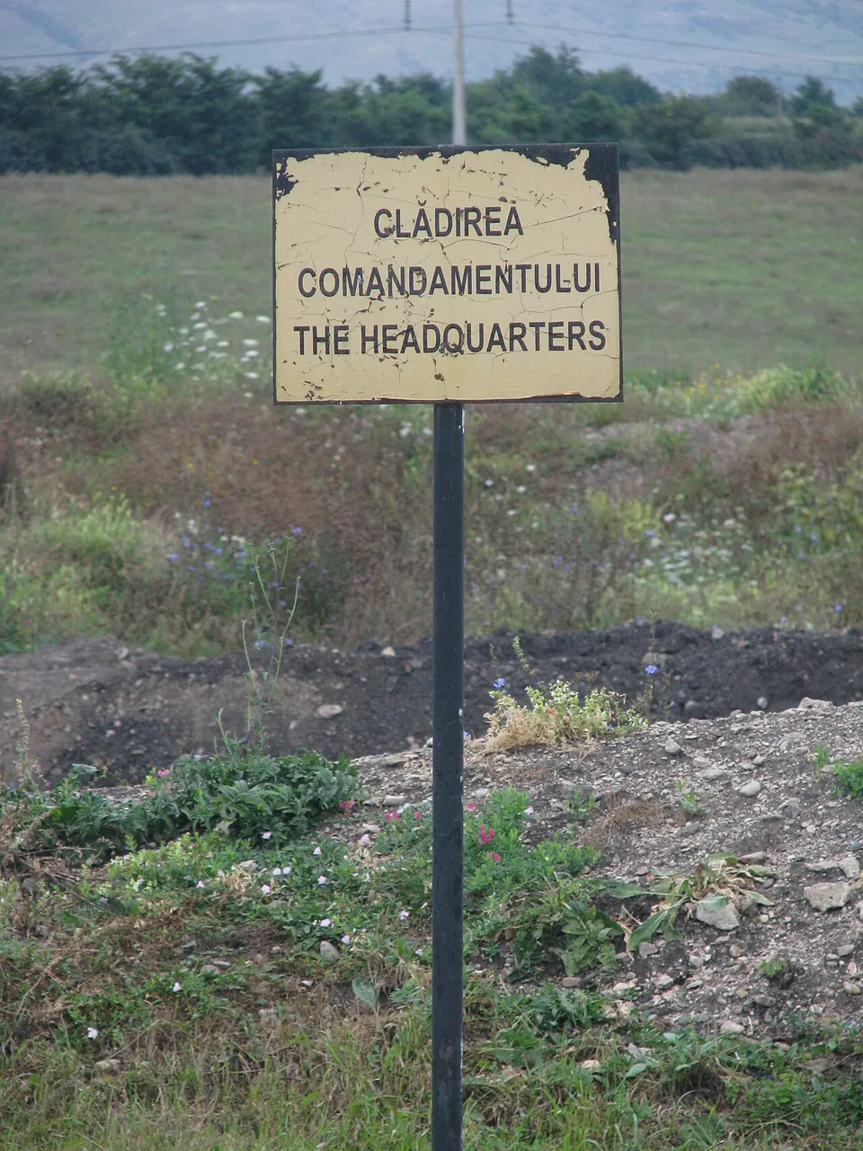 A small weathered yellow sign on a post in rough grass, reading "Clădirea comandamentului – The headquarters".
