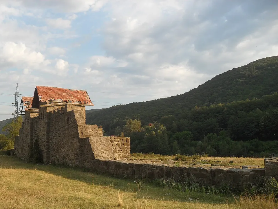 A long stretch of stone wall with a tiled-roof tower at one end, seen across dry grass in evening light, with a wooded hill behind.