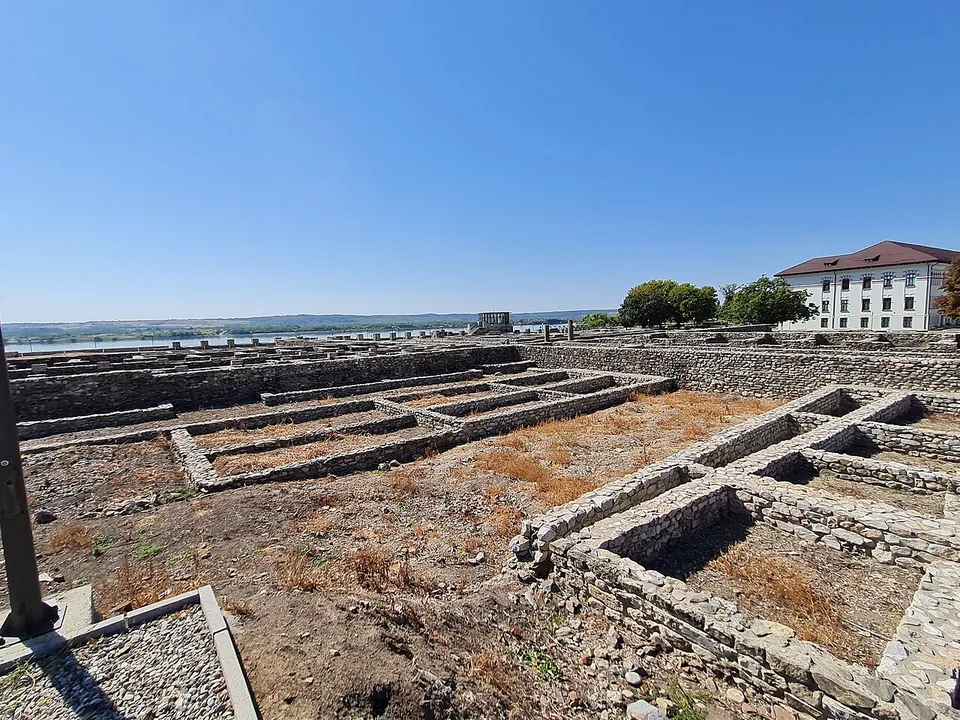 The excavated interior of the fort, rows of low rectangular stone foundations in dry ground, with the wide Danube and its far bank beyond and a white building at the right.