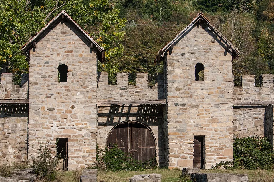 The front of a reconstructed Roman gate, two square stone towers with gabled roofs and small arched windows, joined by a crenellated wall over a wooden double door, with trees behind.