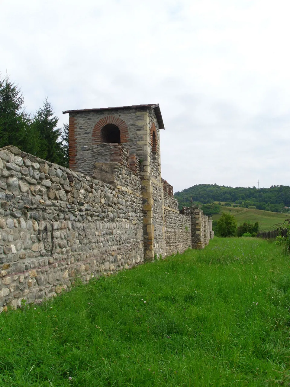 A tall wall of rounded river stones running along a mown bank to a square stone tower with a brick-arched window and a tiled roof, with conifers behind.