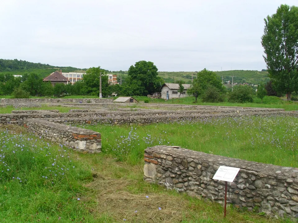 Low walls of river stones outlining buildings in a mown meadow, with a small label on a post, and houses and trees beyond.