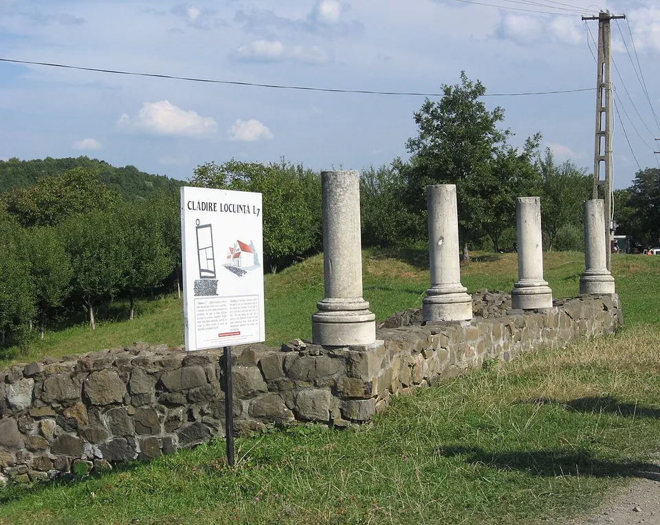 A row of white column shafts standing on a low wall of rough stone beside a grass path, with an information board headed "Clădire locuință L7" in front.