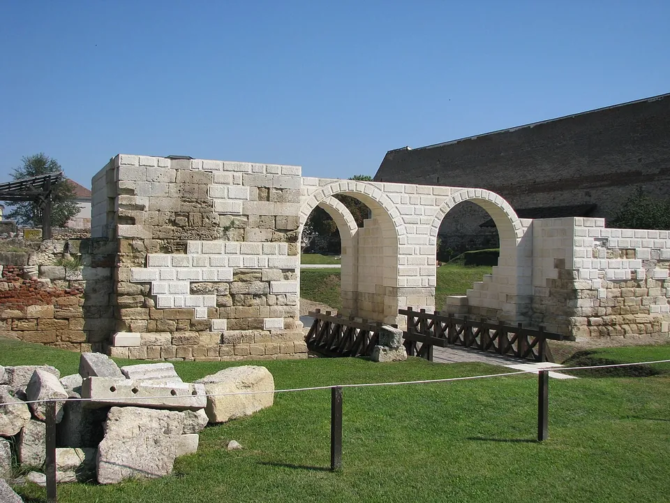 A partly rebuilt Roman gate on a lawn, a block of weathered stone masonry patched with new pale blocks beside two new round arches, with a timber footbridge and loose ancient blocks in front.