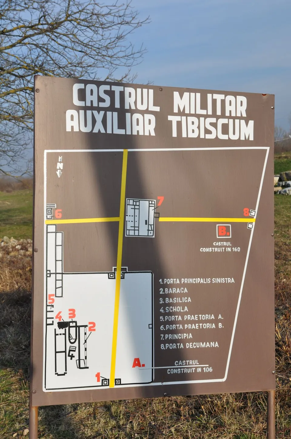 A brown information board headed "Castrul militar auxiliar Tibiscum" with a plan of two forts, one marked as built in 106 and the other in 160, and a numbered key to the gates, barracks, basilica, schola and principia.