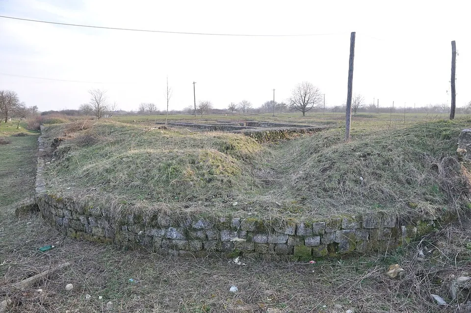 A curving low wall of stone blocks set against a grassy earth bank, with power poles and bare trees on the flat horizon.