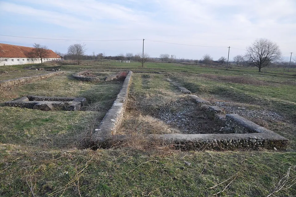 Low consolidated stone foundations of rectangular buildings laid out across a flat grassy field, with a long farm building, bare trees and power lines behind.