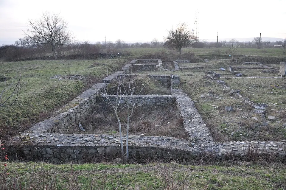 The consolidated stone foundations of a long rectangular building, with a young tree growing inside it and further walls beyond in winter grass.