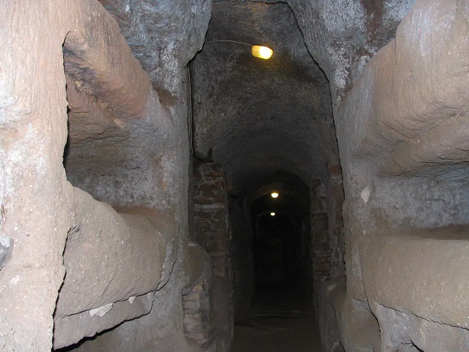 A narrow underground passage cut through pale brown rock, its walls hollowed into rows of empty burial niches, with brick arches and supports further along and small electric lamps lighting the way into the distance.